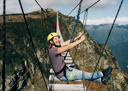 A woman sits on the edge of a rope bridge over a precipice in the mountains
